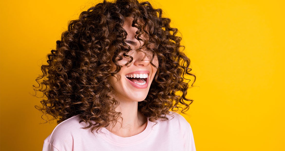 mulher jovem sorri com cabelo cacheado definido, possivelmente, com a técnica dedoliss perfeito